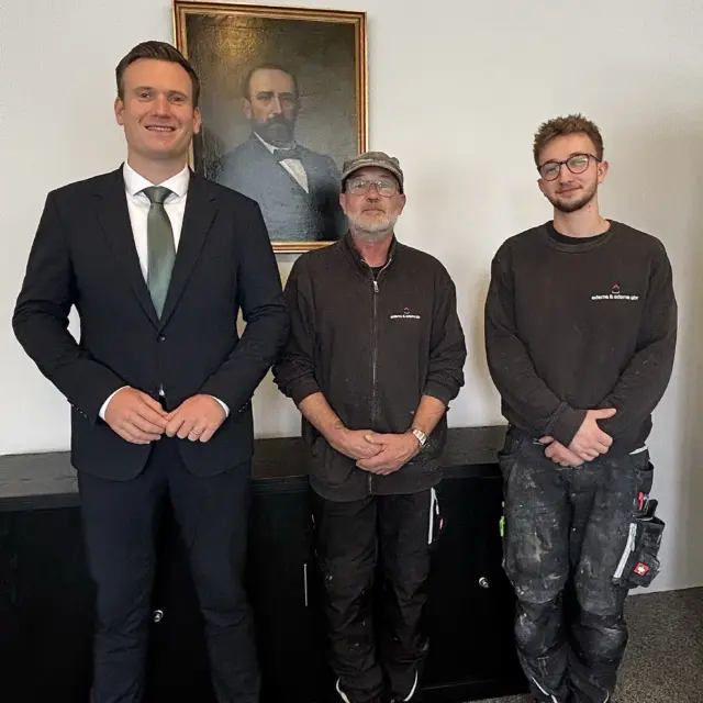 Bürgermeister Philipp Herzog im dunklen Anzug zusammen mit zwei Mitgliedern des Teams der Adams & Adams GbR in Arbeitskleidung, im Hintergrund ein Gemälde
